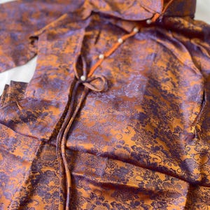 Può includere: Un completo a due pezzi con top con colletto alla coreana e pantaloni abbinati. Il completo è realizzato in un tessuto lucido con un motivo floreale nei toni dell'arancione e del viola. Il top ha una chiusura con fiocco e dettagli di perle.