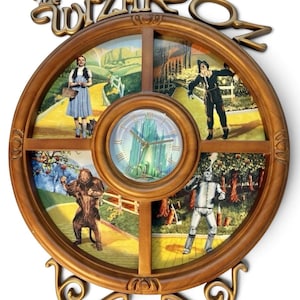 May include: A round wooden wall clock featuring The Wizard of Oz branding at the top. The clock face displays the Emerald City, surrounded by four quadrants depicting Dorothy, the Scarecrow, the Cowardly Lion, and the Tin Man on a yellow brick road.