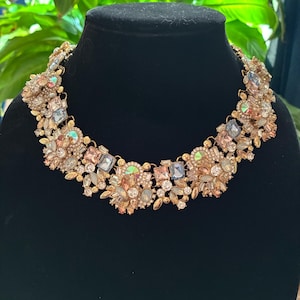 May include: A statement necklace featuring a dense arrangement of multi-shaped crystals and rhinestones in shades of champagne, soft pink, and iridescent blue, set in a gold-tone metal base with a wide, collar-style design and intricate floral clusters.