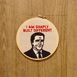 Puede incluir: Bordado en bastidor de madera con la frase "I AM SIMPLY BUILT DIFFERENT" en rojo. La imagen muestra a un hombre sonriente con traje y corbata. El bordado está sobre un fondo de madera.