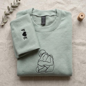 May include: A sage green crewneck sweatshirt featuring black embroidery. The front displays a line art illustration of two figures embracing. The folded sleeve shows the embroidered letters F and E separated by a black heart symbol.