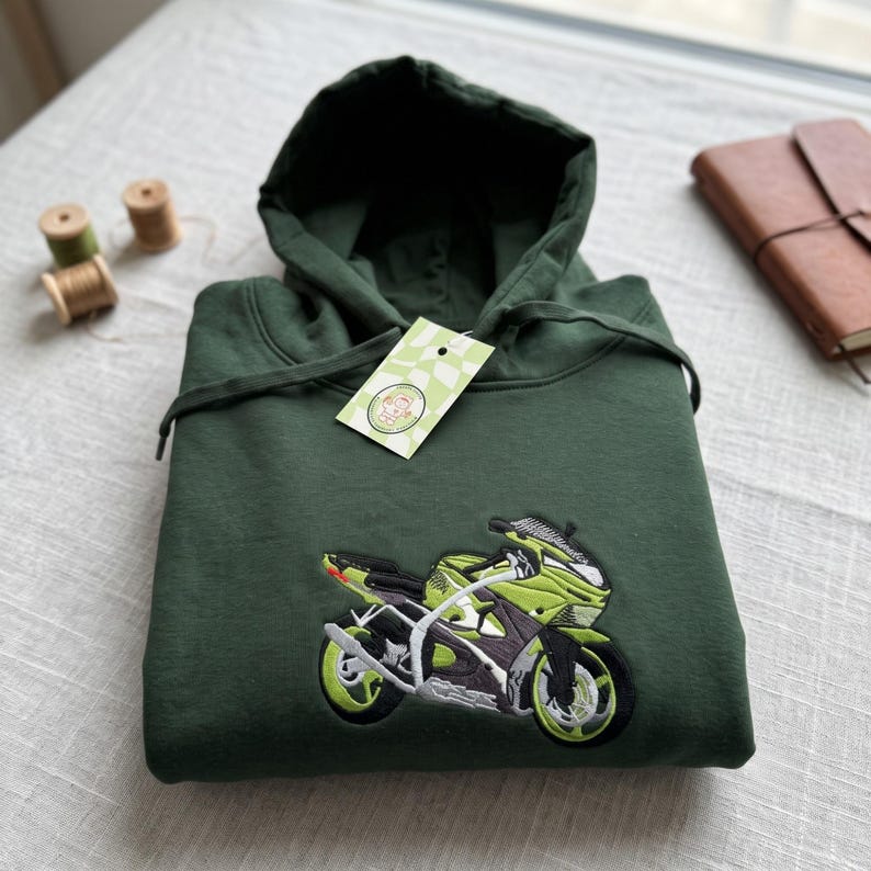 May include: A dark forest green hooded sweatshirt featuring a large, detailed embroidered graphic of a lime green and black sport motorcycle on the front. The hoodie includes matching green drawstrings and a light green tag attached to the neckline.