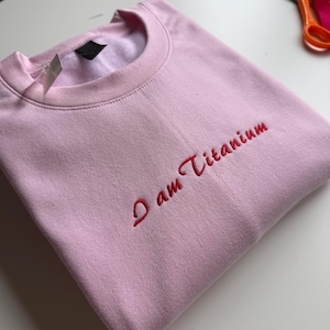 May include: A light pink sweatshirt with the embroidered text "I am Titanium" in red. The crew neck sweatshirt is folded neatly. A tag is attached to the collar.