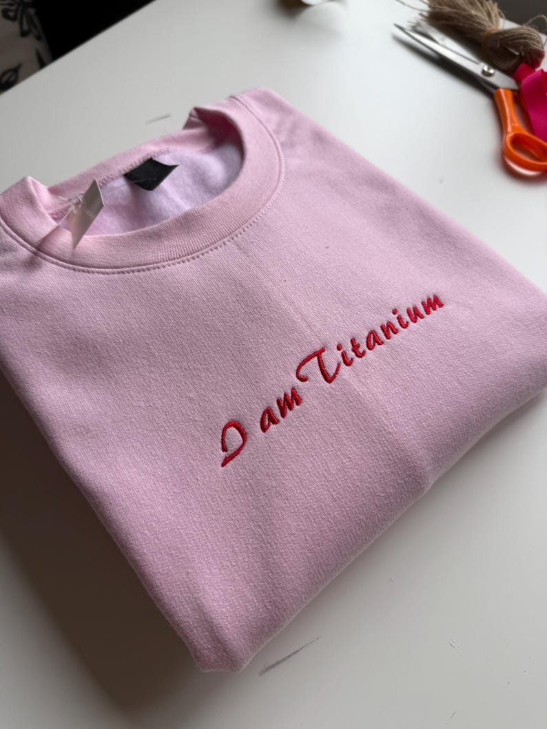 May include: A light pink sweatshirt with the embroidered text "I am Titanium" in red. The crew neck sweatshirt is folded neatly. A tag is attached to the collar.