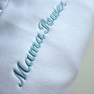 May include: A white sweatshirt with the embroidered text "Mama Power" in a teal script font. The text is vertically aligned on the left side of the garment. The fabric appears soft and comfortable.
