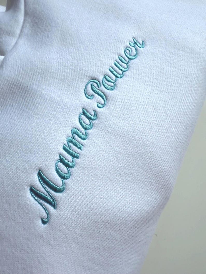 May include: A white sweatshirt with the embroidered text "Mama Power" in a teal script font. The text is vertically aligned on the left side of the garment. The fabric appears soft and comfortable.
