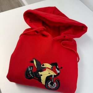 May include: A bright red hooded sweatshirt featuring a detailed embroidered graphic of a yellow and red sport motorcycle on the front center. The hoodie includes a matching red drawstring cord and a large hood, presented in a folded position.