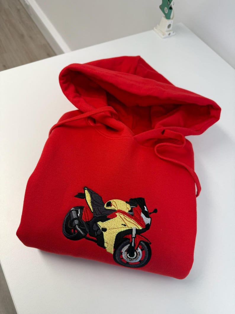 May include: A bright red hooded sweatshirt featuring a detailed embroidered graphic of a yellow and red sport motorcycle on the front center. The hoodie includes a matching red drawstring cord and a large hood, presented in a folded position.