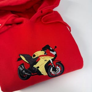 May include: A bright red hooded sweatshirt featuring an embroidered graphic of a sport motorcycle on the front. The motorcycle design is rendered in yellow, red, black, and grey thread, showcasing a detailed profile view of the vehicle.