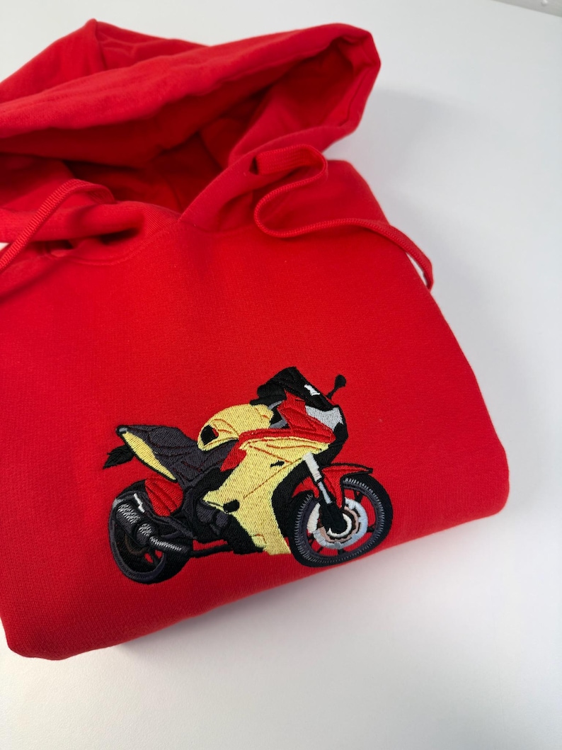 May include: A bright red hooded sweatshirt featuring an embroidered graphic of a sport motorcycle on the front. The motorcycle design is rendered in yellow, red, black, and grey thread, showcasing a detailed profile view of the vehicle.