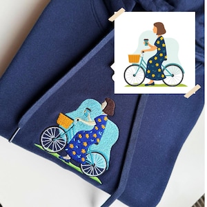 May include: A navy blue hooded sweatshirt featuring an embroidered design of a person with brown hair riding a light blue bicycle. The person wears a dark blue dress with yellow flowers, holds a coffee cup, and has a yellow basket on the bike.