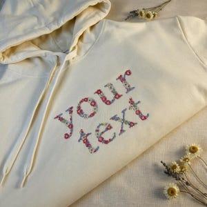 May include: Cream-coloured hoodie with a drawstring hood. The words "your text" are embroidered in a floral pattern using red, blue, and white thread. Dried flowers are scattered around the hoodie.