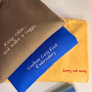 May include: Three sweatshirts in tan, blue, and yellow. The tan sweatshirt features white embroidered text reading Keep calm... and make a cuppa. The blue sweatshirt has white embroidered text reading Custom Cosy Font Embroidery. The yellow sweatshirt has red embroidered text reading Sorry, not sorry.