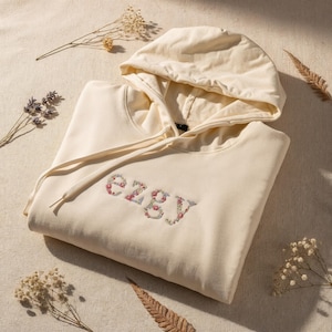 Könnte beinhalten: Cremefarbener Kapuzenpullover mit Kordelzug und dem gestickten Wort "ezgy" in floralem Design. Der Hoodie ist auf einer neutralen Oberfläche gefaltet, mit getrockneten Blumen und Blättern verstreut.