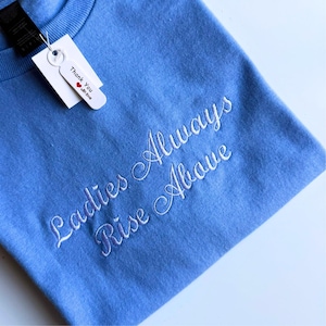 May include: A pale blue crew neck sweatshirt featuring the embroidered phrase "Ladies Always Rise Above" in white cursive. A white tag with the words "Thank You with love" is attached.