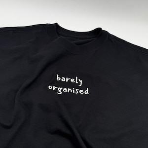 May include: Black crew neck t-shirt featuring the white text "barely organised". The casual t-shirt is made from a soft fabric. The text is in a handwritten style, adding a touch of informality to the garment.