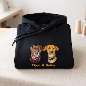 May include: A black hooded sweatshirt featuring an embroidered graphic of two dogs, one brown and one tan, with the text "Pippa & Bobby" stitched in orange thread below the portraits. The hoodie includes a drawstring hood and long sleeves.