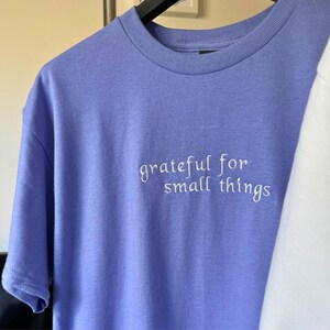 May include: A periwinkle blue t-shirt with the embroidered text "grateful for small things" in white. The short-sleeved shirt is made of a soft material and is hanging on a black hanger.