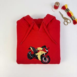 May include: A bright red hooded sweatshirt featuring an embroidered graphic of a yellow and black sport motorcycle on the front. The hoodie includes matching red drawstrings and is displayed alongside a pair of gold-handled scissors and thread.