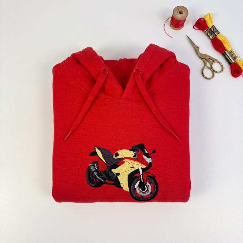 May include: A bright red hooded sweatshirt featuring an embroidered graphic of a yellow and black sport motorcycle on the front. The hoodie includes matching red drawstrings and is displayed alongside a pair of gold-handled scissors and thread.