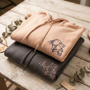 Peut inclure: Deux sweats à capuche superposés, un pêche et un gris anthracite, avec des motifs brodés. Le sweat à capuche pêche a un motif brodé bleu, tandis que le sweat gris présente une broderie couleur pêche. Les deux sweats ont des cordons de serrage.