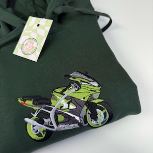 May include: A dark forest green hooded sweatshirt featuring an embroidered graphic of a lime green and black sport motorcycle. A small rectangular tag with a checkered pattern and a circular logo is attached to the hood drawstring.