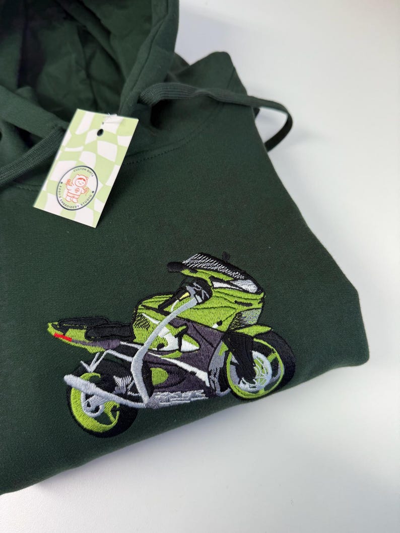May include: A dark forest green hooded sweatshirt featuring an embroidered graphic of a lime green and black sport motorcycle. A small rectangular tag with a checkered pattern and a circular logo is attached to the hood drawstring.