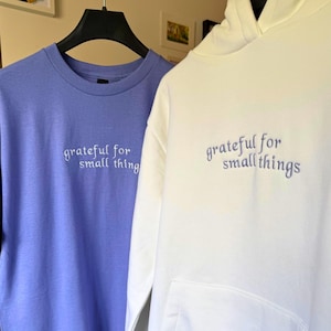 May include: A periwinkle blue crew neck t-shirt and a white hoodie. Both garments have the embroidered text "grateful for small things" in a complementary color. The hoodie has a kangaroo pocket and a drawstring hood.