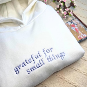 May include: Cream-coloured hoodie with the embroidered text "grateful for small things" in blue. The hoodie has a lined hood and a relaxed fit. A comfortable garment for everyday wear.