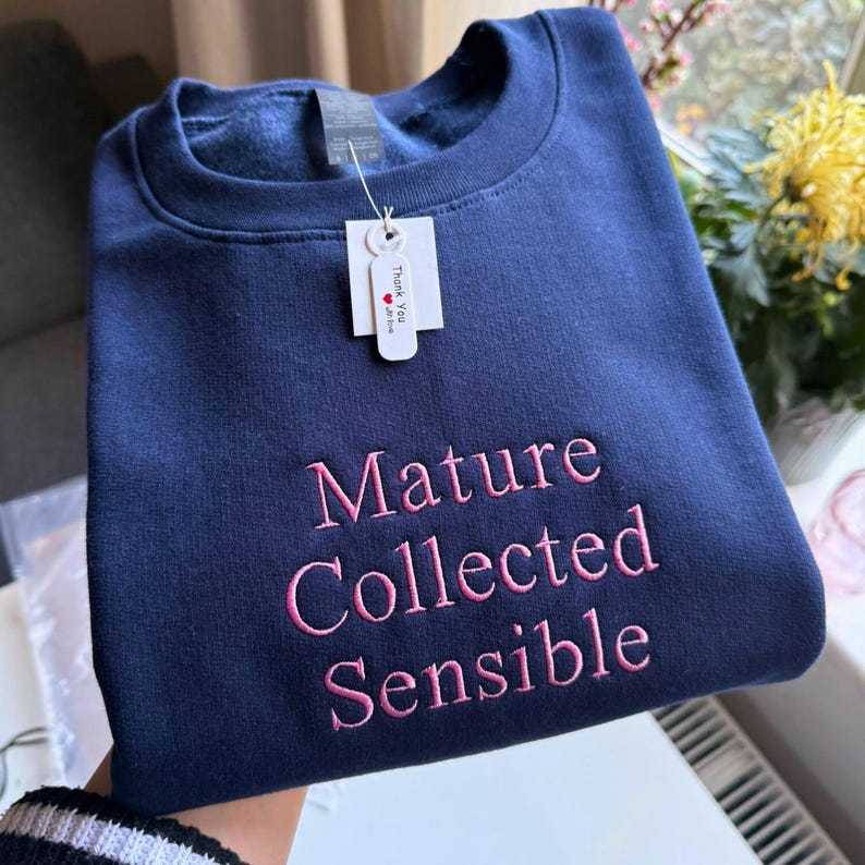 May include: Navy blue sweatshirt with the words "Mature Collected Sensible" embroidered in pink. The sweatshirt has a crew neck and a tag that says "Thank You".