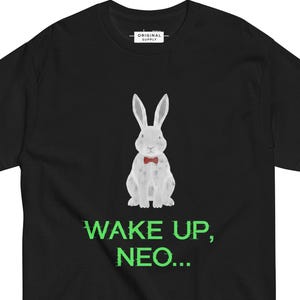 Peut inclure: T-shirt noir avec un lapin blanc portant un nœud papillon rouge. L'expression "WAKE UP, NEO..." est imprimée en vert sous le lapin. Le t-shirt a un col rond et des manches courtes.