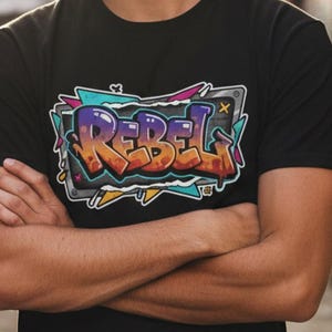 Peut inclure: T-shirt noir avec un motif de style graffiti. Le motif présente le mot "REBEL" en orange, violet et jaune, souligné d'argent, sur fond de formes turquoise et jaunes. La personne porte le t-shirt avec les bras croisés.