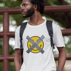 May include: White t-shirt with a yellow smiley face design, crossed with grey tape. The smiley has black eyes and a curved mouth. The shirt is worn by a person with a black backpack.