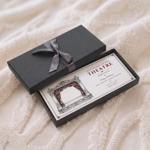 May include: A black rectangular gift box with a matching satin ribbon bow containing a printed ticket. The ticket features a detailed illustration of a theatre stage with red curtains and text reading AN EVENING AT THE THEATRE FOR LUCY.