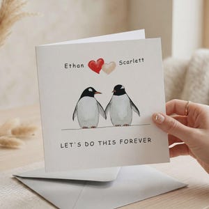 Puede incluir: Tarjeta cuadrada con una ilustración de dos pingüinos tomados de la mano. Los nombres "Ethan" y "Scarlett" están encima, con dos corazones. El texto "LET'S DO THIS FOREVER" está escrito debajo. La tarjeta es sostenida por una mano.