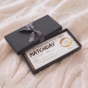 May include: A black rectangular gift box with a dark gray ribbon, containing a white novelty ticket for a matchday event. The ticket features the text Get Ready For Matchday, a gold scratch-off circle, and details for a 21st birthday gift.