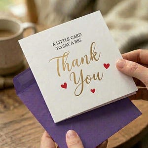 Puede incluir: Una tarjeta de felicitación blanca con la inscripción dorada "Thank You" y "A little card to say a big". La tarjeta tiene tres pequeños acentos de corazón rojo y se sostiene sobre un sobre morado.
