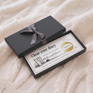Puede incluir: Una caja de regalo rectangular negra con una cinta de satén gris, que contiene una tarjeta de rascar blanca. La tarjeta presenta el texto "Clear your diary", "A proper day out awaits" y "Cricket day for two" en un área circular dorada.