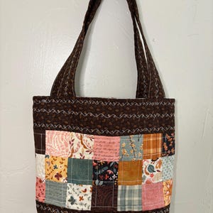 Könnte beinhalten: Eine braune Tragetasche mit Patchwork-Quadraten in verschiedenen Farben und Mustern. Die Tasche hat eine einfarbige braune Basis und Griffe mit einem dekorativen, genähten Rand. Der Patchwork umfasst Blumen-, Karo- und Textdruckstoffe.