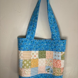 Puede incluir: Un bolso tote hecho a mano con tela azul estampada con flores y cuadrados de patchwork de varios colores. El bolso tiene correas largas y forma rectangular.