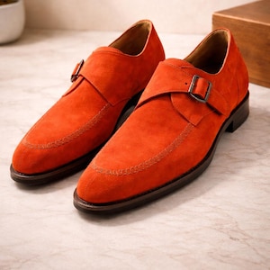 Könnte beinhalten: Ein Paar orangefarbene Monk-Strap-Schuhe aus Wildleder mit Ziernähten an der Spitze und einem Schnallenverschluss. Die Schuhe haben eine dunkelbraune Sohle und ein Lederfutter. Sie sind auf einer Marmoroberfläche ausgestellt.