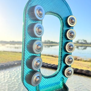 Puede incluir: Un objeto en forma de U de color turquesa con una superficie texturizada y múltiples detalles circulares plateados. El objeto está situado al aire libre con un fondo borroso de agua y cielo.