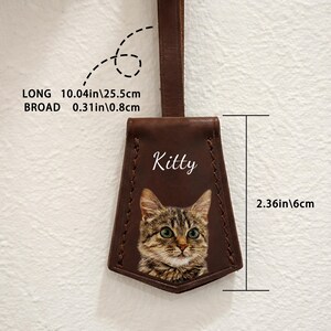 May include: A brown leather tag with a cat portrait and the name "Kitty" in white script. The tag is 10.04 inches long and 0.31 inches broad. The cat's image is detailed with green eyes and brown and grey fur. The tag is 2.36 inches wide.