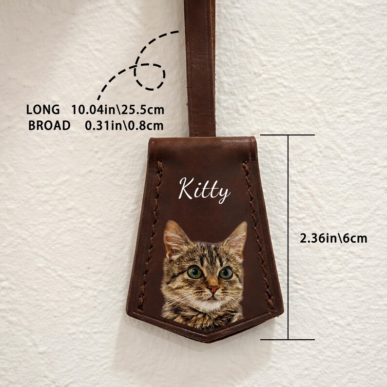 May include: A brown leather tag with a cat portrait and the name "Kitty" in white script. The tag is 10.04 inches long and 0.31 inches broad. The cat's image is detailed with green eyes and brown and grey fur. The tag is 2.36 inches wide.