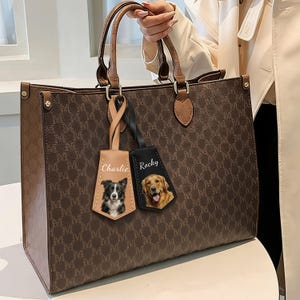 May include: Brown tote bag with a repeating "M" pattern. The bag has brown handles and trim. Two personalized dog tags hang from the handle, one tan with a black and white dog and the name "Charlie", and one black with a golden retriever and the name "Rocky".