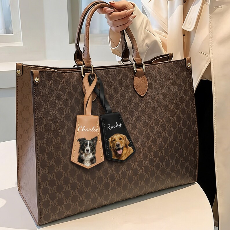 May include: Brown tote bag with a repeating "M" pattern. The bag has brown handles and trim. Two personalized dog tags hang from the handle, one tan with a black and white dog and the name "Charlie", and one black with a golden retriever and the name "Rocky".