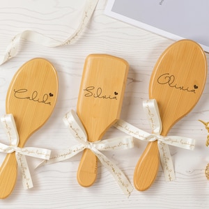 May include: Three wooden hairbrushes with personalized names: Calida, Silvia, and Olivia. Each brush is tied with a white ribbon. The names are written in a cursive font, with a small heart symbol next to each name.