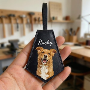 May include: Black leather luggage tag with a photo of a dog and the name "Rocky" in white script. The dog is brown and white with its mouth open. The tag has a black loop at the top.