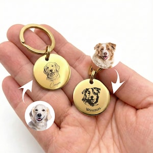 May include: Two gold-colored, round keychains with engraved dog portraits. One keychain has the name "Cooper" below the dog's image, and the other has "Winston." The keychains are held in a hand, with additional dog photos shown.