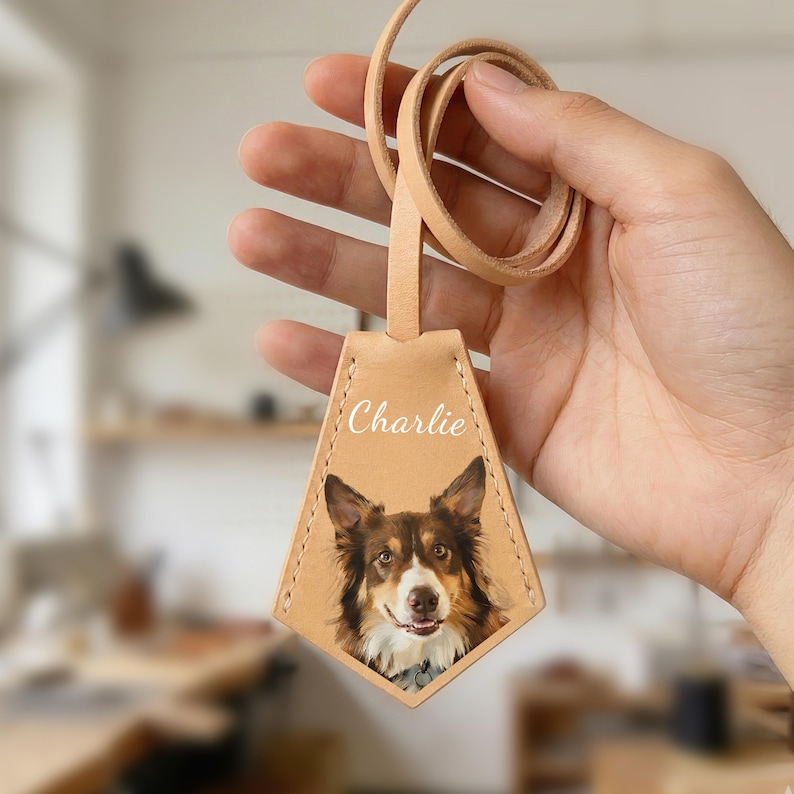 May include: A light tan leather dog tag with a photo of a brown and white dog. The tag has the name "Charlie" printed on it. The tag is attached to a matching leather loop.